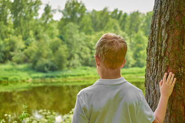 A European teenager stands near the river and holds on to a tree with his hand