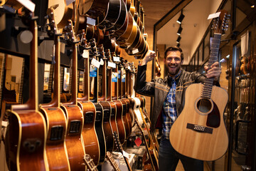 Portrait of talented caucasian musician in leather jacket who has just purchased classic acoustic guitar in music shop. - obrazy, fototapety, plakaty