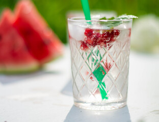 drink with pomegranate berries, green currant leaves and ice cubes, on a white wooden background, watermelon slices in the background, sunny day.
