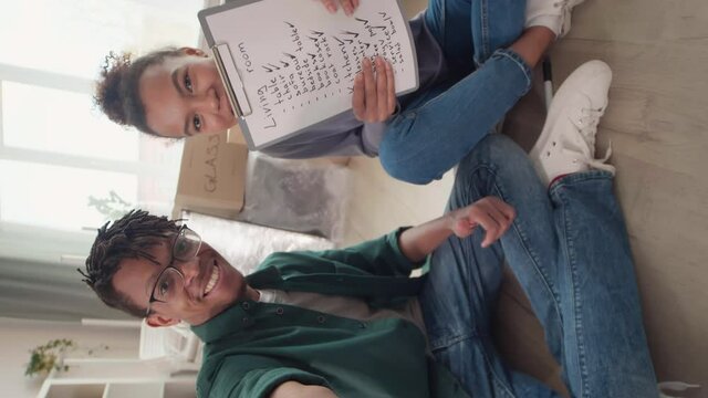 Vertical view shot of young African-American man and his girlfriend sitting together on floor in living room and talking to friend by video link while getting ready for move