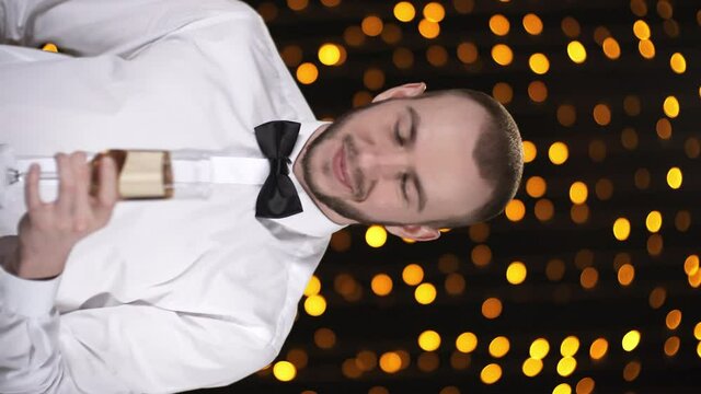 Vertical portrait shot of young man in bowtie standing in dark room with fairy lights and raising his glass of champagne while looking at camera, then drinking