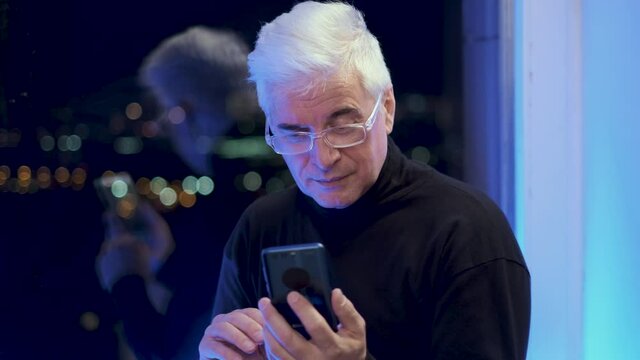 Pensive old retired gentleman in turtleneck sweater searching and scrolling via smartphone. Elder graying businessman by the window. Grey haired male person. Senior gaffer man with white shaved face.