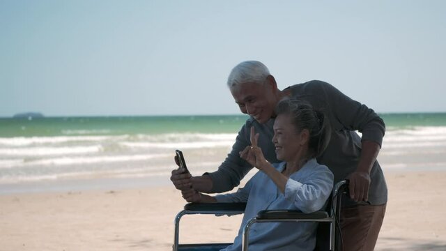 Happy Asian elderly woman sitting in wheelchair and husband is a wheelchair user smartphone taking selfie on the beach, summer vacation, Retirement couple concept