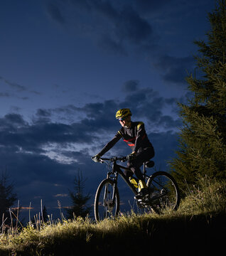 Smiling young man riding bicycle downhill with beautiful blue evening sky on background. Bicyclist in sports cycling suit cycling down grassy hill at night. Concept of sport, biking and active leisure