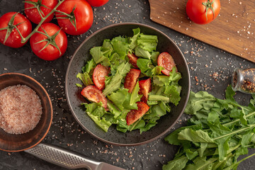 There is a bowl of light summer vegetable salad of tomatoes and greens and fresh ingredients for its cooking on a black background. Top view