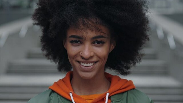 Portrait of young attractive toothy smiling woman looking at the camera while standing at city center. Closeup face of beautiful woman with curly hair. Woman appearance concept.