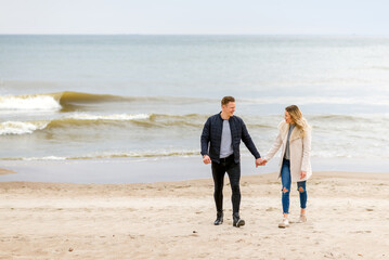 Attractive young couple walking along the shore of a sandy beach, on a spring romantic holiday, outdoors.