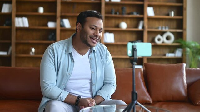 Blogging concept. Young indian male blogger in casual clothes streaming video online or recording tutorial for social media network, looking at the mobile phone on a tripod and laughing