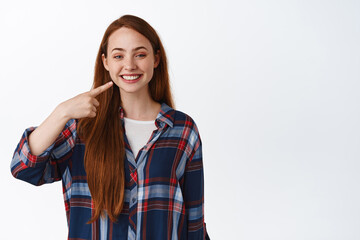 Whitening and stomatology. Smiling redhead teen girl pointing at her perfect white teeth, recommend dental clinic, standing in plaid shirt against white background