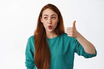 Surprised and impressed ginger girl, say wow, super cool news, looking left at logo and thumb up in...