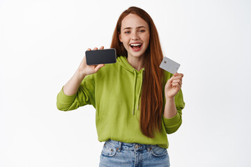 Online shopping. Attractive modern ginger girl makes purchased, paying in internet, showing credit card and horizontal smartphone screen, white background