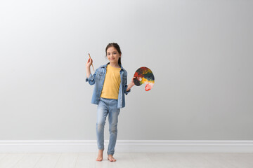 Little girl with brush and paint palette near light grey wall indoors - Powered by Adobe