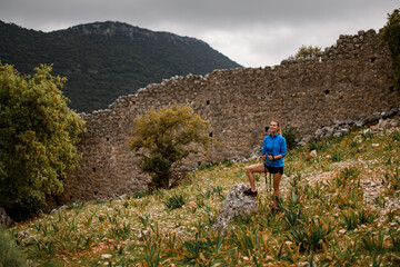Fototapeta premium young smiling woman with trekking poles against the background of stone wall and mountain