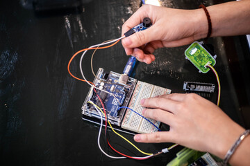 A close-up of hands working on an Arduino Uno connected to a breadboard with jumper wires. One hand...