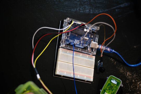 A close-up of an Arduino Uno connected to a breadboard with jumper wires. One hand holds a small module—possibly a sensor or buzzer—while the other hand steadies the setup during assembly.