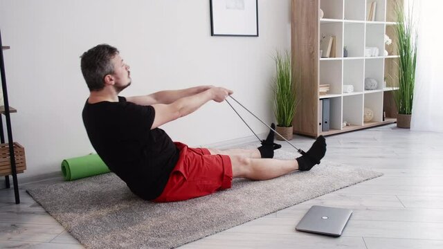 Home training. Funny man. Fitness beginner. Healthy sport. Middle-aged chubby guy sitting floor closing laptop cover playing fool with resistance band in light room interior.