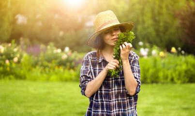 A young girl gardener in a straw hat holds a bouquet of harvested fresh mint and inhales its wonderful menthol scent, a woman is harvesting in the garden. - Powered by Adobe