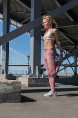 Attractive sporty woman goes in for sports outdoors under the bridge on sunny day. Female sportsman warming up and stretch her legs before run and yoga practice