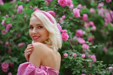 Obraz na płótnie Canvas Happy smiling beautiful blonde woman wearing pink headband posing in blooming rose garden. Copy, empty space for text