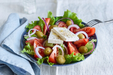 Greek salad of fresh vegetable with tomatoes, lettuce, olives, red onion and feta cheese in bowl on white striped linen tablecloth. Selective focus