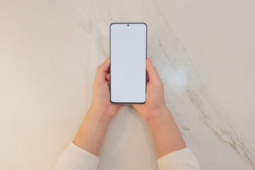 top view of hands holding blank white screen mock up mobile phone with white marble background