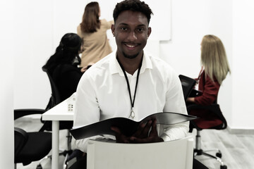 Confident team leader. African young man holding office binder and looking at camera with smile while his colleagues working in the background.