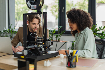 serious interracial colleagues sitting near 3D printer, laptop and plastic figures in modern office