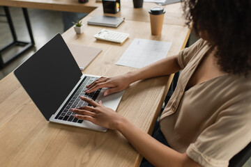 partial view of young african american woman sitting at desk with paper cup and typing on laptop in modern office