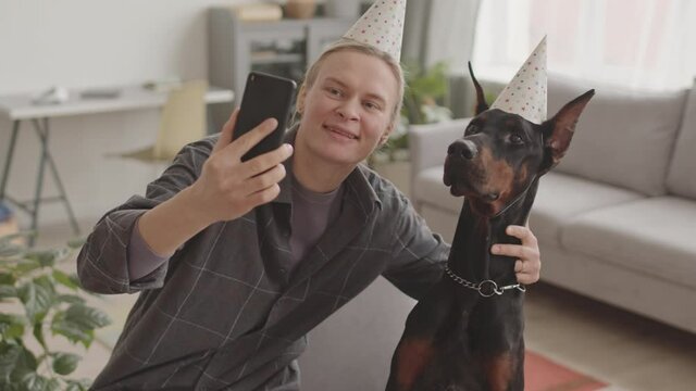 High angle medium long of young Caucasian woman and black Doberman dog wearing party hats, taking selfie pictures at home