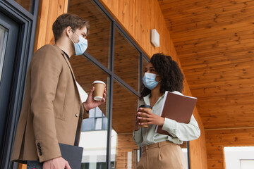 young interracial colleagues in medical masks talking with coffee paper cups on street near building