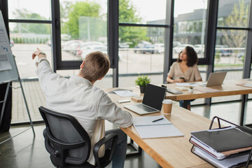 young man pointing with finger at flip chart near blurred african american colleague in modern office