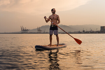 Young man paddle surfs in the sea at sunset