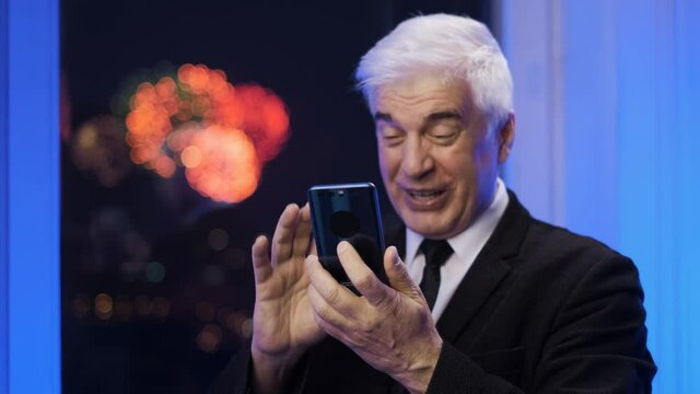 Smiling happy old retired gentleman in blazer suit looking at smartphone. Elder graying businessman by the window with fireworks. Grey haired male person. Senior gaffer man with white shaved face.