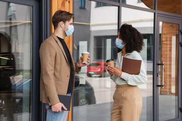 young interracial colleagues in medical masks talking with paper cups in hands on street near building
