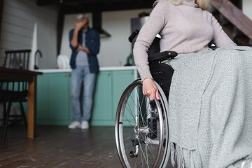 Fototapeta na wymiar Senior woman sitting in wheelchair near husband in kitchen on blurred background