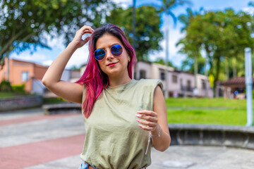 medium shot of a woman in a park touching her hair with her hands, wearing blue sunglasses and with fuchsia hair