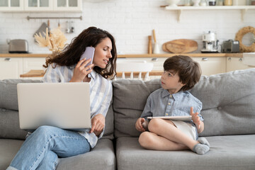 Fototapeta na wymiar Multitasking casual businesswoman and mother work from home talk to kid, make phone call use laptop sit on couch in kitchen. Modern mom remote worker or freelancer lifestyle during covid-19 lockdown