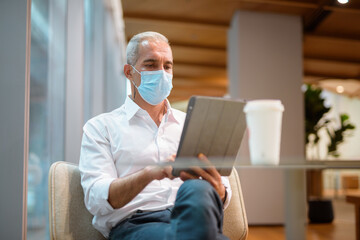 Businessman sitting at coffee shop and using digital tablet while wearing face mask and social distancing