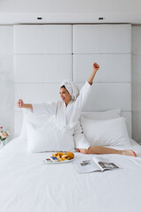 Woman in bathrobe sitting in bed, having breakfast and taking selfie at hotel room