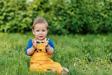 Happy charming baby boy in fashionable clothes sitting on the grass in the garden on a summer day