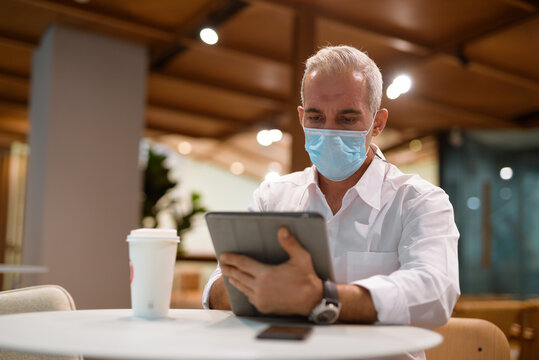 Businessman sitting at coffee shop and using digital tablet while wearing face mask and social distancing