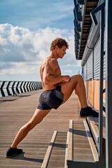 Fototapeta na wymiar Young muscular tattooed man doing exercises, stretching early in the morning on pier by the sea