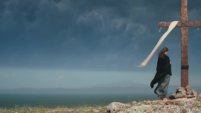 A woman in a black cape approaches the Holy Cross on a stormy day. Pan shot