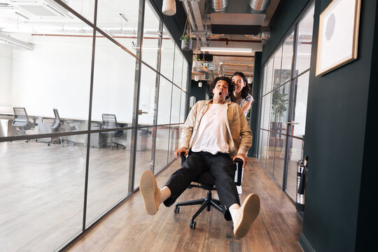 Young woman having fun pushing man on chair along walkway in open plan office