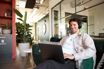 Young man online with mobile phone and headphones relaxing in seating area of open plan office