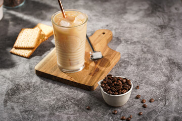 A glass with ice cubes and milk coffee on a wooden board, with brass straw and coffee beans