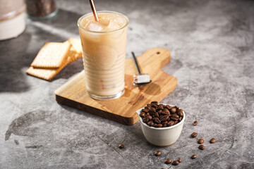 A glass with ice cubes and milk coffee on a wooden board, with brass straw and coffee beans