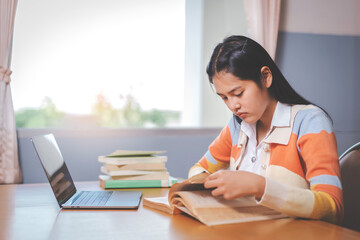 Young Asian woman college student in student uniform studying, reading a book, laptop in university or college library. Youth girl student and tutoring education with a technological learning concept