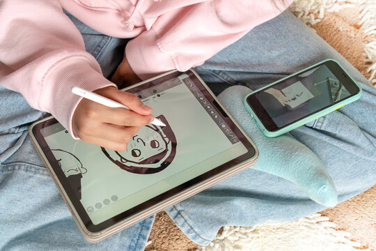 Focused stylish teenage girl painting using Apple Pencil on IPad tablet and watching online course on phone while practicing at home. Online training, online classes. Russia, Tatarstan, June 15, 2021
