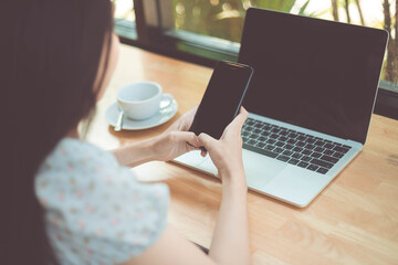 Woman hand holding white mobile phone on a table with a laptop in office.

